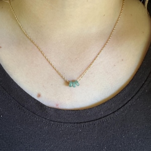 May include: A delicate gold-colored necklace featuring three small, light green gemstones. The necklace is worn against a black top, highlighting the contrast between the gold chain and the gemstones.
