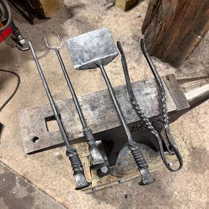 May include: A set of five handcrafted, dark metal blacksmithing tools, including a poker, fork, shovel, and tongs, arranged on an anvil. The tools have twisted handles and are designed for working with fire and metal.