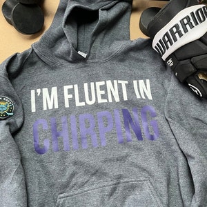 May include: A gray hooded sweatshirt with the text "I'm fluent in chirping" in white and purple lettering. The sweatshirt has a pocket on the front and a small patch on the left sleeve.