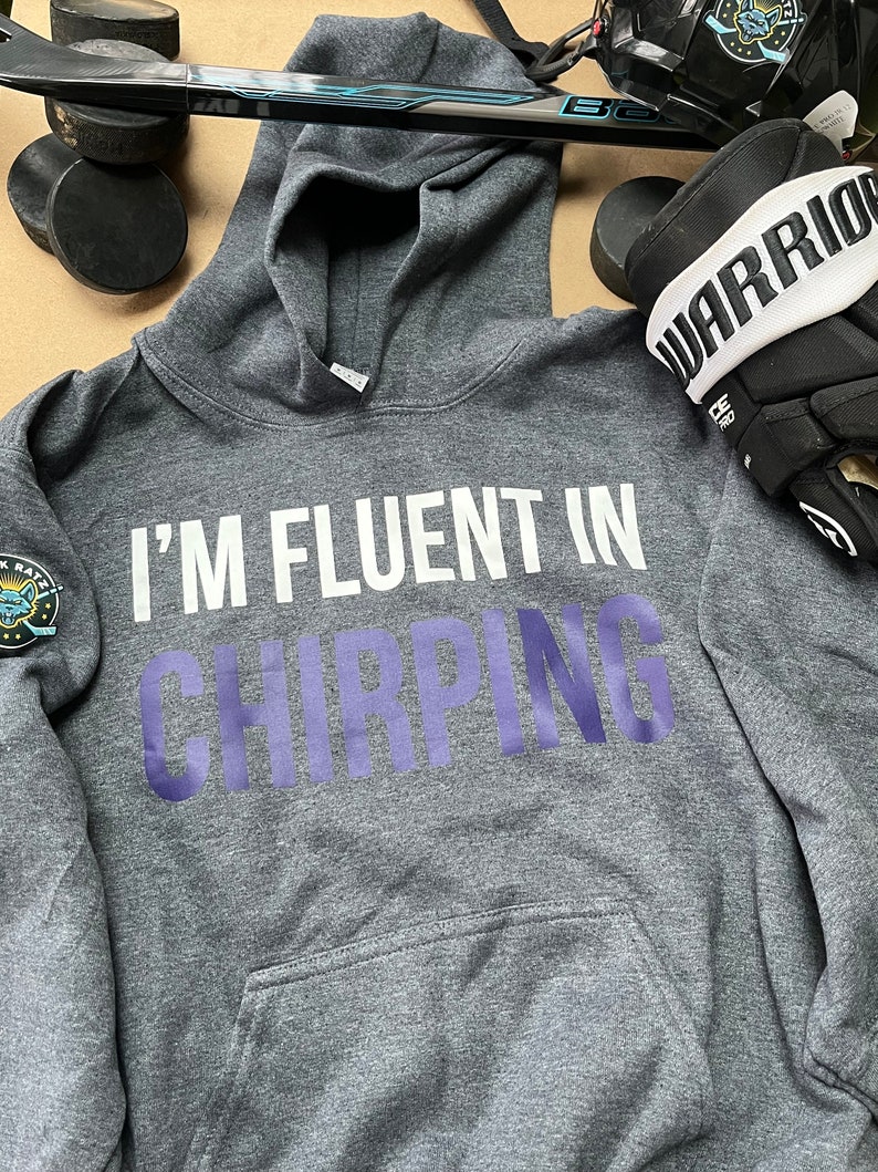 May include: A gray hooded sweatshirt with the text "I'm fluent in chirping" in white and purple lettering. The sweatshirt has a pocket on the front and a small patch on the left sleeve.