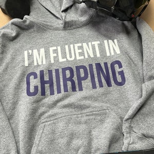 May include: Gray hooded sweatshirt with the text "I'm fluent in chirping" in white and navy blue lettering. The sweatshirt is lying on a surface with a hockey stick and a black hockey helmet.