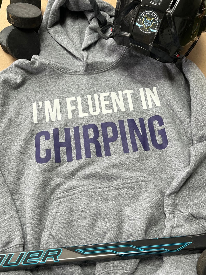 May include: Gray hooded sweatshirt with the text "I'm fluent in chirping" in white and navy blue lettering. The sweatshirt is lying on a surface with a hockey stick and a black hockey helmet.