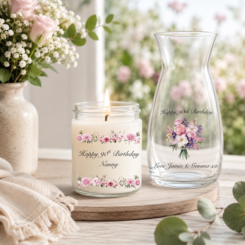 May include: A glass candle jar with a lit wick and a clear glass vase, both featuring floral graphics and text. The candle label reads Happy 90th Birthday Nanny, while the vase is inscribed with Happy 90th Birthday and Love James & Gemma xx.