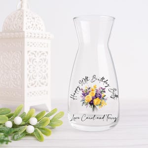 Pode incluir: Uma jarra de &aacute;gua de vidro transparente com um design floral de flores amarelas e roxas. O texto diz "Happy 90th Birthday Betty" e "Love Carol and Terry". Uma lanterna decorativa branca est&aacute; no fundo.