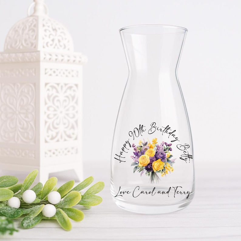 Pode incluir: Uma jarra de &aacute;gua de vidro transparente com um design floral de flores amarelas e roxas. O texto diz "Happy 90th Birthday Betty" e "Love Carol and Terry". Uma lanterna decorativa branca est&aacute; no fundo.