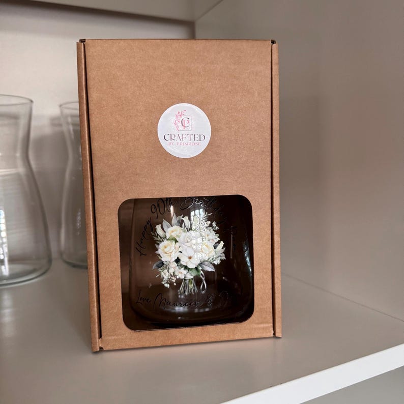 Pode incluir: Um copo transparente com um design floral e o texto "Happy 90th Birthday, Love Momma" &eacute; exibido dentro de uma caixa de papel&atilde;o marrom. O copo apresenta um buqu&ecirc; de flores brancas. A caixa tem um adesivo circular com o texto "Crafted by Crimsone".