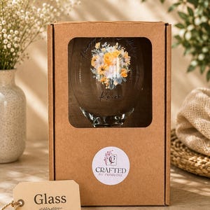 May include: A clear glass inside a brown cardboard box with a rectangular cutout. The glass features a floral bouquet print in yellow and white, the text "Happy Birthday" and "Karen", and a round sticker reading "CRAFTED BY PRIMROSE". A beige tag labeled "Glass" is attached.