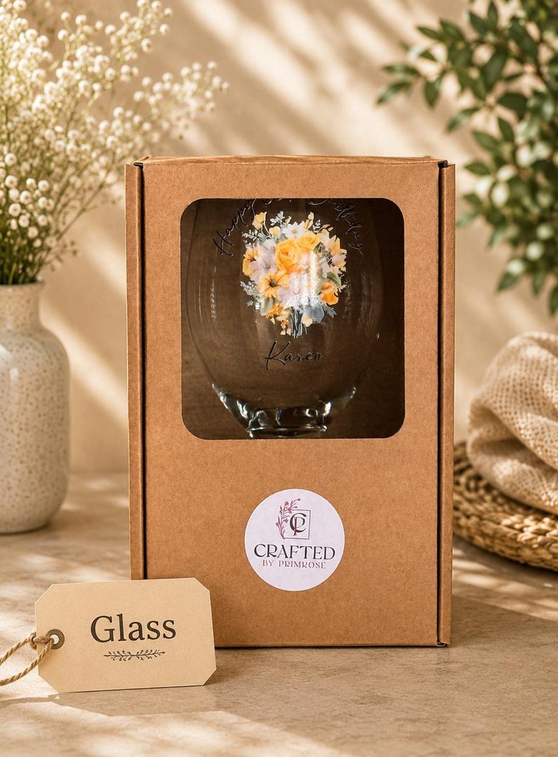 May include: A clear glass inside a brown cardboard box with a rectangular cutout. The glass features a floral bouquet print in yellow and white, the text "Happy Birthday" and "Karen", and a round sticker reading "CRAFTED BY PRIMROSE". A beige tag labeled "Glass" is attached.