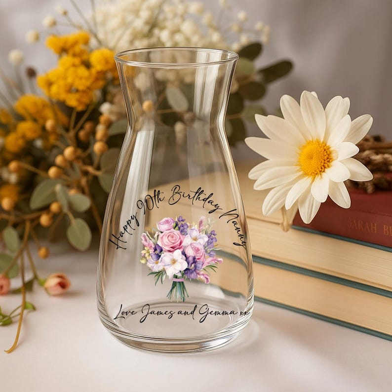Pode incluir: Um vaso de vidro transparente com um design de buqu&ecirc; floral e o texto "Happy 90th Birthday Nanny, Love James and Gemma xx". O vaso &eacute; cercado por flores e livros.