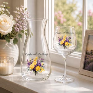 May include: A clear glass carafe and a matching wine glass, both featuring a printed bouquet of yellow and purple flowers and the text Happy 18th Birthday in black script. The glassware is transparent with a smooth, curved design.