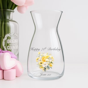 Pode incluir: Um vaso de vidro transparente com base larga e pesco&ccedil;o estreito, com o texto "Happy 70th Birthday" e "Jean xx". Um buqu&ecirc; floral de flores amarelas e brancas &eacute; impresso no vaso. O vaso est&aacute; vazio.