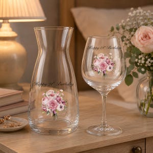 May include: A clear glass carafe and a matching wine glass, both featuring a printed bouquet of pink roses and the text Happy 21st Birthday. The carafe has a tapered neck and a wide base, while the wine glass stands on a slender stem.