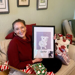 May include: A woman is holding a framed illustration of a dog with the name "Hector" written below it. The illustration is in black and white and the dog is sitting with its tongue sticking out. The woman is sitting on a couch with a white throw blanket and a red pillow behind her. There are wrapped gifts on the couch next to her.