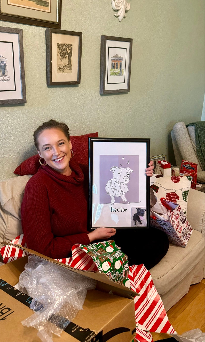 May include: A woman is holding a framed illustration of a dog with the name "Hector" written below it. The illustration is in black and white and the dog is sitting with its tongue sticking out. The woman is sitting on a couch with a white throw blanket and a red pillow behind her. There are wrapped gifts on the couch next to her.