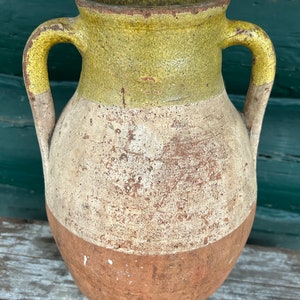 May include: A large, terracotta pottery vase with two handles. The vase is glazed in a yellow, white, and brown color scheme. The vase is weathered and shows signs of age.