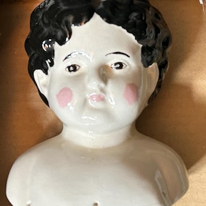 May include: A ceramic doll head and shoulder bust featuring a glossy white finish, black painted hair with a curly texture, brown eyes, and prominent pink circular blush on the cheeks. The facial features are defined with painted details.