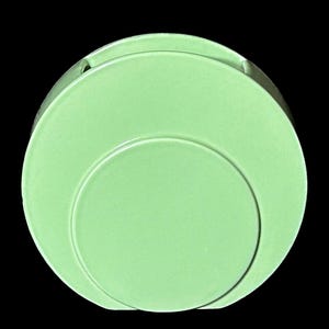May include: A light green ceramic ashtray with a circular design. The ashtray has a recessed area for holding cigarettes and a smooth, glossy finish. The design features a smaller circle within a larger one.