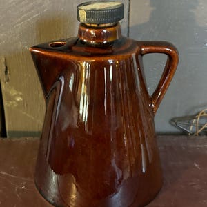 May include: A brown ceramic syrup pitcher with a handle and a spout. The pitcher has a dark brown lid and a glossy finish. The design is a classic, vintage style, suitable for serving maple syrup or other condiments.