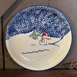 May include: A round, white ceramic plate with a blue rim and a winter scene. The plate features a snowman wearing a red hat and scarf, a green tree, and a blue dotted sky. The plate measures approximately 10 inches in diameter.