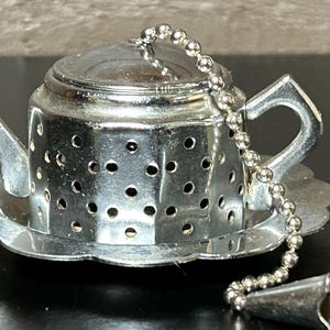 May include: A silver-colored tea infuser shaped like a teapot, with a matching saucer and a chain with a flower-shaped end. The infuser has small holes and a handle. The teapot infuser is designed for steeping loose leaf tea.