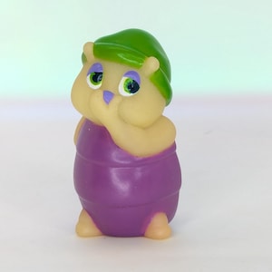 May include: A small, whimsical figurine of a cartoon character. The figure has a light yellow body, a purple outfit, and a green hat. The eyes are large with green irises and purple accents. The character is holding a finger to its mouth.