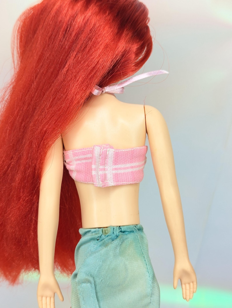 Op de afbeelding: Een pop met lang, levendig rood haar vastgebonden met een roze lint. De pop draagt een roze en wit gestreepte top en een lichtblauwe rok. De huid van de pop is lichtbruin.