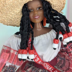 May include: A doll wearing a straw hat and a red and white plaid dress. The doll has long black braids adorned with red and white beads. A red ribbon across the chest reads "NATIONAL COSTUME".