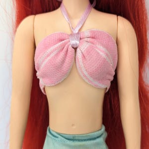 Op de afbeelding: Een pop met rood haar en een roze top met witte strepen. De top is met een roze lint om de nek gebonden. De pop draagt een lichtblauwe rok.
