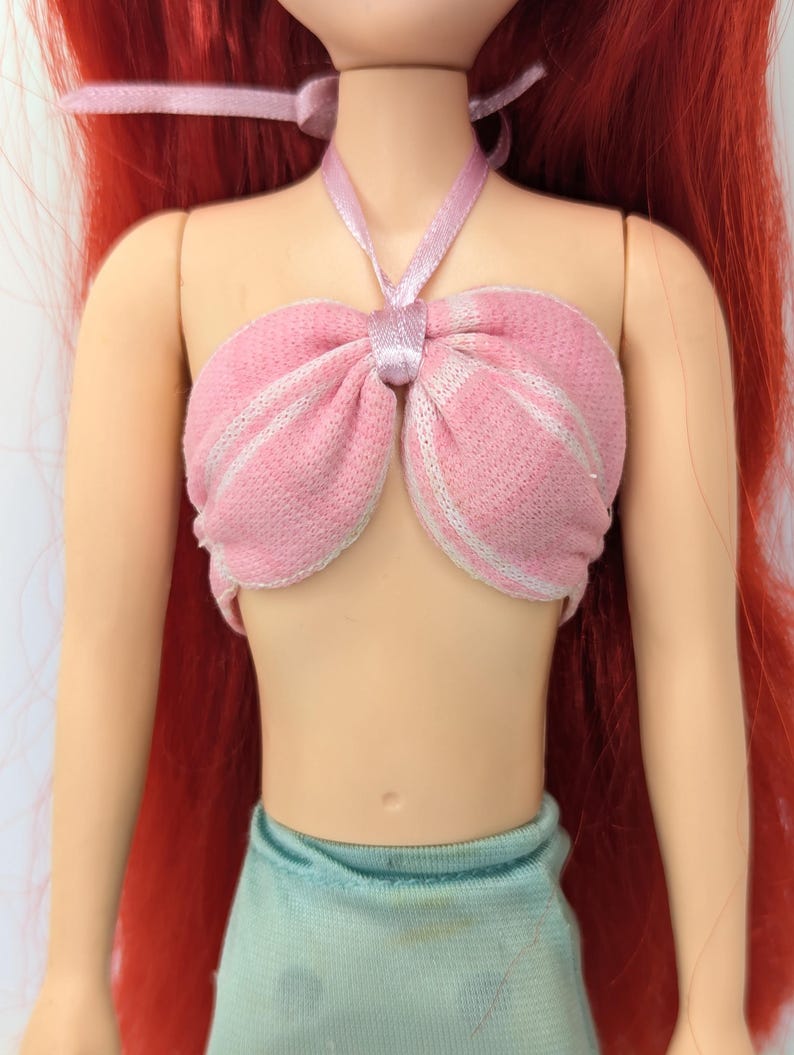 Op de afbeelding: Een pop met rood haar en een roze top met witte strepen. De top is met een roze lint om de nek gebonden. De pop draagt een lichtblauwe rok.