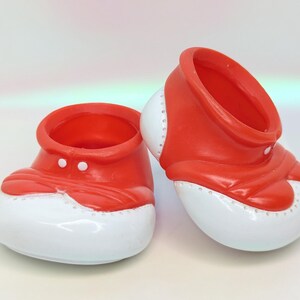May include: A pair of red and white decorative baby shoes. The shoes have a red upper with a rounded white toe and a small white button detail. The shoes are designed to be decorative.