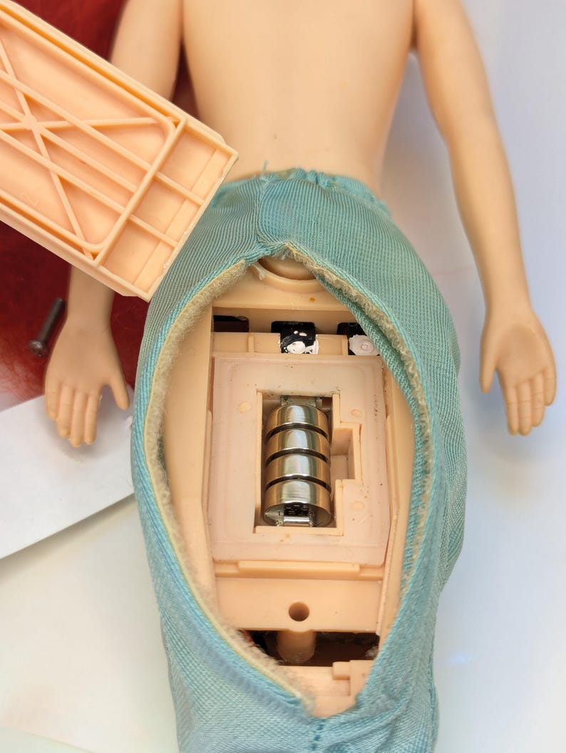 Op de afbeelding: Een pop met een turquoise rok, die de interne componenten onthult. De rug van de pop is open en toont een batterijcompartiment met vier zilveren batterijen. Een licht perzik plastic onderdeel wordt verwijderd.