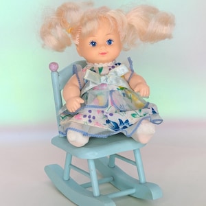 May include: A vintage doll with blonde pigtails, blue eyes, and a floral dress sits on a light blue wooden rocking chair. The doll's dress features a delicate floral pattern with blue, pink, and white accents. The chair has a rounded back and curved rockers.