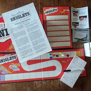 May include: A vintage board game titled "The Game of Sniglets" with a red and white color scheme. The game includes a game board, instruction manual, score sheets, and game pieces. The box reads "The Sni for people who didn't."