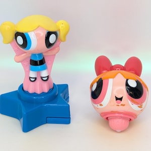 May include: Two Powerpuff Girls figurines. One is a standing figure with a yellow head, pink dress, and blue base. The other is a spinning top with a red bow and pink face. Both have large eyes.