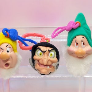 May include: Three Disney character keychains: a winking dwarf in a yellow hat, a wicked witch, and a smiling dwarf in a green hat. Each keychain has a pink or blue clip and a white beard. The background is a gradient of pink and white.