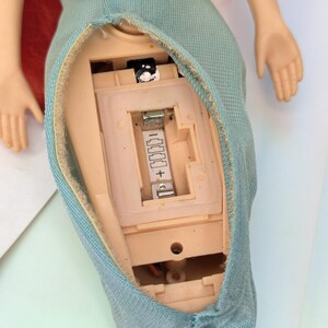 Op de afbeelding: Een pop met rood haar en een lichtblauwe rok. De torso van de pop is open en toont het interne batterijcompartiment en de elektronische componenten. De armen van de pop zijn uitgestrekt en de handen zijn zichtbaar.