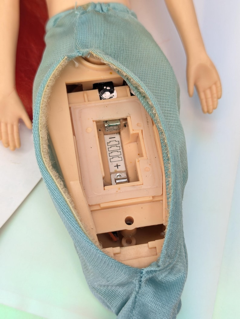 Op de afbeelding: Een pop met rood haar en een lichtblauwe rok. De torso van de pop is open en toont het interne batterijcompartiment en de elektronische componenten. De armen van de pop zijn uitgestrekt en de handen zijn zichtbaar.