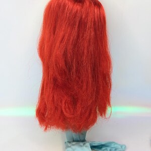 Op de afbeelding: Een poppenhoofd met lang, levendig rood haar. Het haar is gestyled in een vloeiende, gelaagde manier. De nek van de pop en de aangehechte kleding zijn lichtblauw van kleur. De achtergrond is wit.