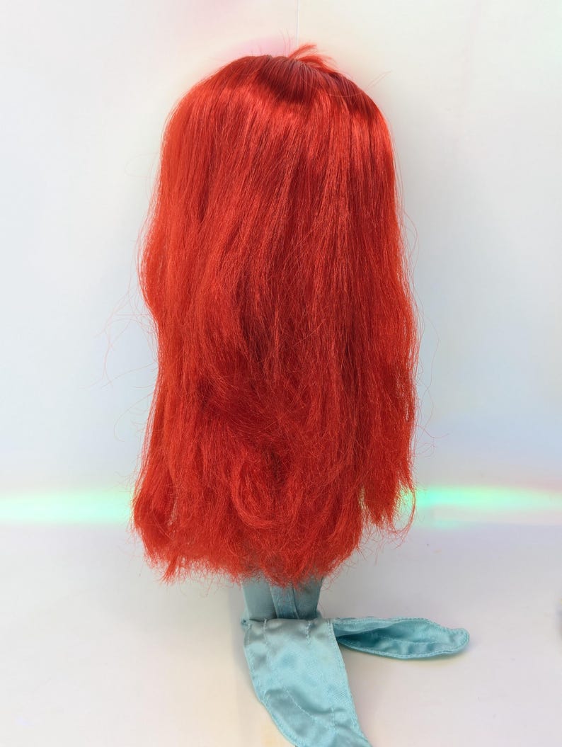 Op de afbeelding: Een poppenhoofd met lang, levendig rood haar. Het haar is gestyled in een vloeiende, gelaagde manier. De nek van de pop en de aangehechte kleding zijn lichtblauw van kleur. De achtergrond is wit.