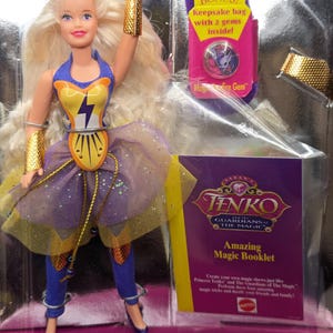 May include: A purple and yellow doll with blonde hair wearing a purple and yellow outfit. The doll is holding a gold bracelet in her right hand. The doll is in a plastic package with a purple and yellow background. The package also includes a booklet titled "Amazing Magic Booklet".