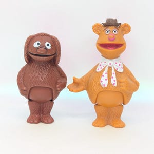 May include: Two vintage Muppet Show character figurines. One is a brown, shaggy dog-like character with large white eyes. The other is Fozzie Bear, an orange bear wearing a hat and bow tie with pink polka dots. Both are standing upright.