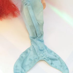 Op de afbeelding: Een pop met rood haar en een lichtblauwe zeemeerminstaart. De staart is gemaakt van een zijdeachtige stof en heeft een vinachtige vorm. De torso en arm van de pop zijn zichtbaar.