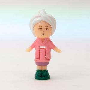 May include: A small plastic figurine of a person wearing a white towel on their head and a pink robe. The figurine has a light skin tone, purple skirt, and green shoes. The arms are outstretched.