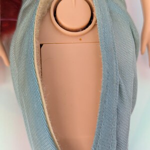 Op de afbeelding: Close-up van de torso van een pop met een lichtblauwe denim-achtige stoffen rok. Het lichaam van de pop is zichtbaar door een opening in de rok, waar een beige plastic component met een cirkelvormig ontwerp te zien is. De rok heeft een klittenbandsluiting.