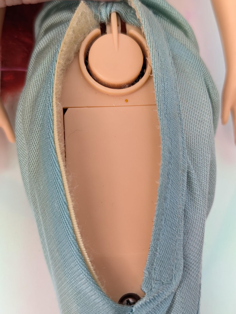 Op de afbeelding: Close-up van de torso van een pop met een lichtblauwe denim-achtige stoffen rok. Het lichaam van de pop is zichtbaar door een opening in de rok, waar een beige plastic component met een cirkelvormig ontwerp te zien is. De rok heeft een klittenbandsluiting.