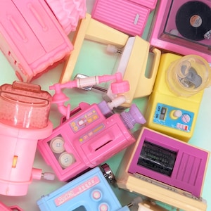 May include: A collection of miniature plastic toy appliances and furniture including a pink wardrobe, a yellow blender, a blue sewing machine, a pink video camera, a record player, and a television with text reading Barbie Mattel Skipper.