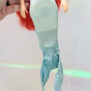 Op de afbeelding: Een pop met rood haar en een lichtblauwe zeemeerminstaart. De staart van de pop is gemaakt van een zachte, vloeiende stof. De pop wordt door een hand vastgehouden.
