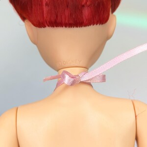 Op de afbeelding: Een pop met rood haar en een roze lint om haar nek. De achterkant van de pop is zichtbaar, met het Disney-logo. De pop draagt een roze en witte jurk. De huid van de pop heeft een licht perzikkleur.