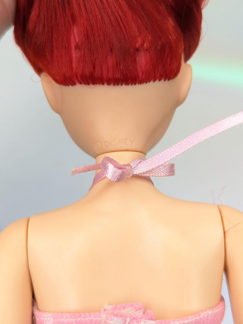 Op de afbeelding: Een pop met rood haar en een roze lint om haar nek. De achterkant van de pop is zichtbaar, met het Disney-logo. De pop draagt een roze en witte jurk. De huid van de pop heeft een licht perzikkleur.