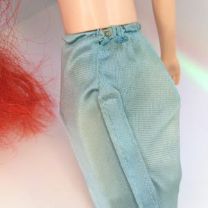 Op de afbeelding: Een pop met een roze en wit gestreept topje en een lichtblauwe broek. De pop heeft rood haar en een lichte huidskleur. De broek heeft een kleine knoopsluiting in de taille en een verticale naad.